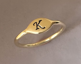 May include: A gold ring with a small oval-shaped top featuring the letter "K" engraved in black.