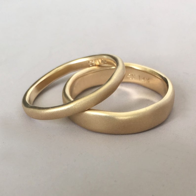 Op de afbeelding: Twee gouden trouwringen met een geborstelde afwerking. De ringen zijn op elkaar gestapeld. De bovenste ring heeft de inscriptie "585 14K".