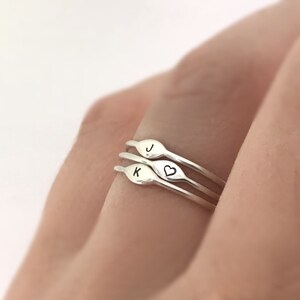 May include: Three silver rings stacked on a finger. The top ring has the letter "J" engraved on it. The middle ring has the letter "K" engraved on it and a small heart on the side. The bottom ring is plain.