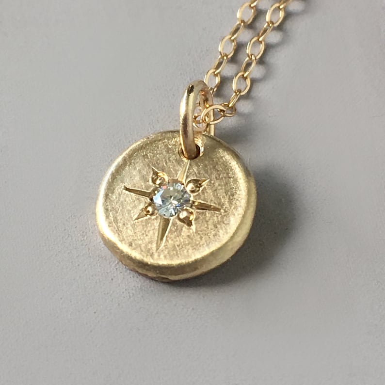 May include: A gold-colored pendant necklace with a round, textured charm. The charm features a starburst design with a small, light blue gemstone at its center. The chain is delicate and also gold-toned.