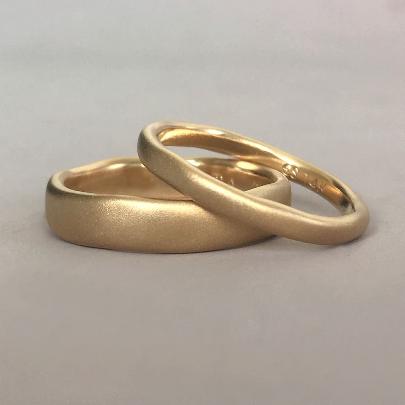 Op de afbeelding: Twee gouden trouwringen, waarvan er &eacute;&eacute;n iets breder is dan de andere, met een geborstelde matte afwerking.