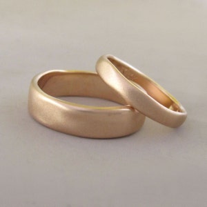 Op de afbeelding: Twee eenvoudige, geborstelde gouden trouwringen. De ringen zijn glad en hebben een afgeronde vorm.