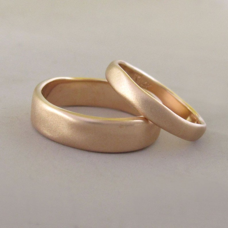 Op de afbeelding: Twee eenvoudige, geborstelde gouden trouwringen. De ringen zijn glad en hebben een afgeronde vorm.