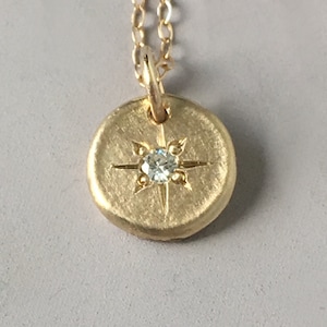 May include: A gold-colored pendant necklace featuring a circular charm with a starburst design and a small, clear gemstone at the center. The pendant hangs from a delicate gold chain, creating a simple yet elegant piece of jewelry.
