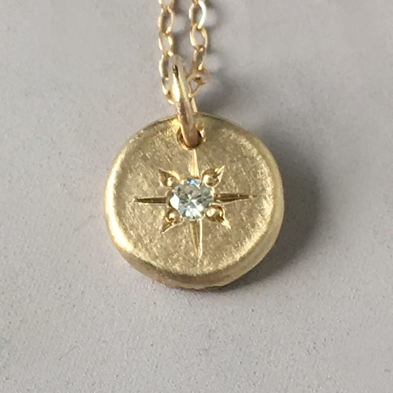 May include: A gold-colored pendant necklace featuring a circular charm with a starburst design and a small, clear gemstone at the center. The pendant hangs from a delicate gold chain, creating a simple yet elegant piece of jewelry.
