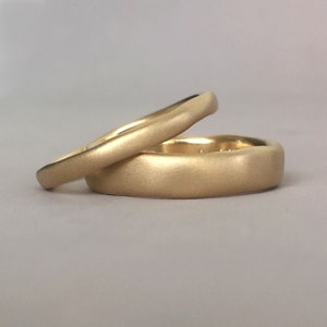 Op de afbeelding: Twee gouden trouwringen met een geborstelde afwerking. De ringen zijn op elkaar gestapeld.