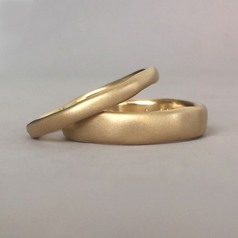 Op de afbeelding: Twee gouden trouwringen met een geborstelde afwerking. De ringen zijn op elkaar gestapeld.