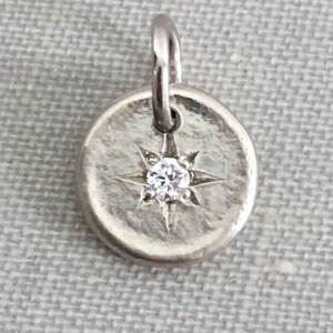 May include: A silver charm with a small diamond set in the center of a starburst design.