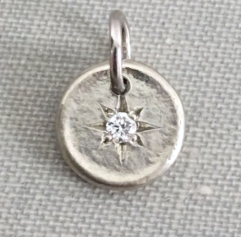 May include: A silver charm with a small diamond set in the center of a starburst design.