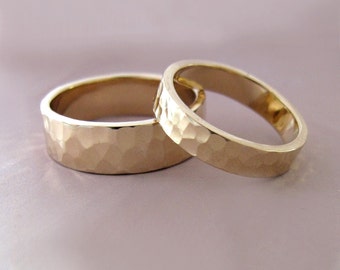May include: Two gold wedding bands with a hammered texture. The rings are side-by-side, showcasing their simple, elegant design. The bands have a flat profile and a warm, golden hue, perfect for a wedding or anniversary.
