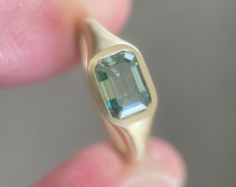 May include: A gold ring with a rectangular, emerald-cut, green gemstone. The ring has a smooth, polished band and a bezel setting that surrounds the gemstone. The gemstone has a slight blue hue.