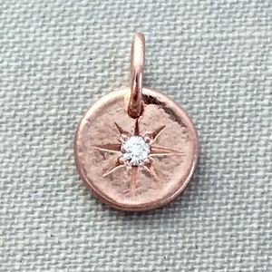 May include: A rose gold charm with a small diamond in the center of a starburst design.