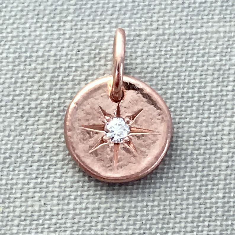 May include: A rose gold charm with a small diamond in the center of a starburst design.