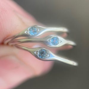 May include: A stack of three silver rings, each featuring a light blue gemstone at the center of an elongated, eye-shaped design. Small, clear accent stones flank the central gem, adding sparkle. The rings are stacked, creating a layered effect.
