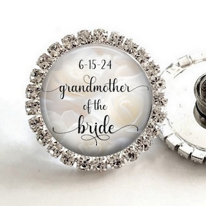 May include: A silver pin with a white background and the text "6-15-24 grandmother of the bride" in black cursive lettering. The pin is surrounded by clear rhinestones.