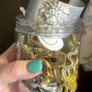May include: A clear jar filled with various jewelry pieces. A silver cuff bracelet with a lion's head design is at the top. Other items include gold necklaces, rings, and a white, curved object. The jar is held by a hand with teal nail polish.