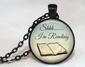 Può includere: Una collana con ciondolo rotondo dotata di una cornice e una catena in metallo nero. L'inserto centrale mostra l'illustrazione di un libro aperto su sfondo crema con il testo nero Shhh....... I'm Reading in carattere corsivo e serif.