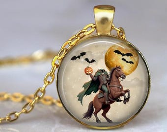 May include: Gold-toned pendant necklace featuring a round charm with a Halloween-themed illustration. The charm depicts a headless figure on horseback, holding a pumpkin, with bats and a full moon in the background. The chain is gold.