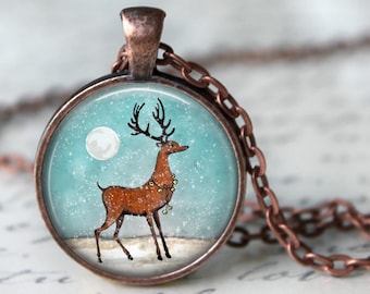 Op de afbeelding: Een ronde hangerketting met een koperkleurige ketting. De hanger toont een winterse scène met een bruin rendier, een volle maan en vallende sneeuw. Het rendier heeft een slinger van belletjes om zijn nek.