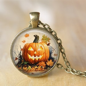 May include: A round pendant necklace with a bronze chain. The pendant features a smiling jack-o'-lantern with autumn leaves. The pumpkin is orange with glowing yellow eyes and mouth. The background includes bare branches and a tombstone.