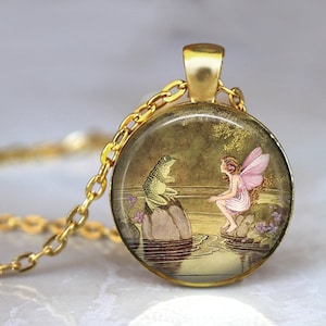 Puede incluir: Un collar de oro con un colgante redondo de vidrio que presenta una hada con alas rosadas sentada en una roca junto a un estanque, hablando con una rana verde sentada en una roca.