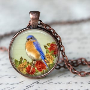 May include: A round pendant necklace with a copper finish. The pendant features a blue and orange bird perched on a branch, surrounded by red and yellow roses. The bird is set against a light green background.
