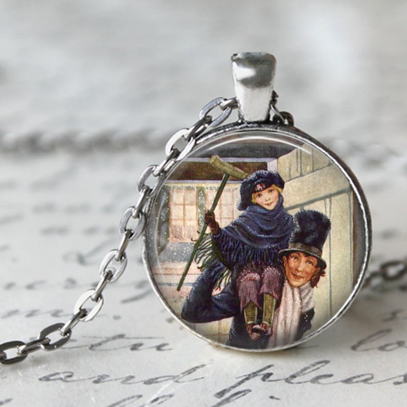 K&ouml;nnte beinhalten: Eine silberfarbene Halskette mit Anh&auml;nger, die einen runden Charm mit einer Vintage-Illustration zeigt. Die Illustration zeigt zwei Figuren in Winterkleidung, von denen eine die andere tr&auml;gt. Der Charm h&auml;ngt an einer feinen Kette.