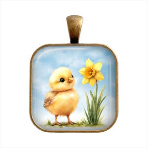 May include: A yellow chick with a fluffy body and black eyes looks at a yellow daffodil with a green stem and leaves against a blue sky background. The image is on a square glass pendant with a bronze frame.
