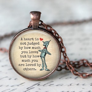 Puede incluir: Un collar de cobre con un colgante redondo de vidrio que presenta una cita sobre el amor y una imagen de dibujos animados del Hombre de Hojalata de El Mago de Oz. La cita dice: "Un corazón no se juzga por cuánto amas; sino por cuánto eres amado por los demás."