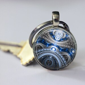May include: A round keychain with a silver metal ring and a decorative pendant. The pendant features a blue, black, and silver abstract design. A key is visible in the background.