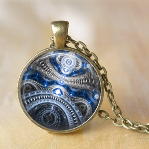 May include: A round pendant necklace with a bronze-colored chain. The pendant features a blue, white, and black abstract design. The design has a mechanical or futuristic aesthetic. The necklace is displayed on a neutral background.