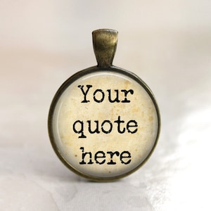 Puede incluir: Un colgante redondo de oro antiguo con un inserto de vidrio. El inserto de vidrio tiene un fondo de papel vintage envejecido con el texto "Your quote here" impreso en negro.
