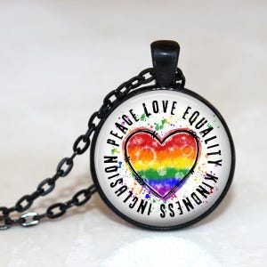 May include: A round pendant necklace with a black chain. The pendant features a rainbow heart design with the words "Peace, Love, Equality, Kindness, Inclusion" around the heart. The heart has a tie-dye effect.