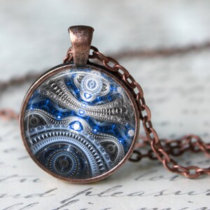 May include: A round pendant necklace with a copper-colored chain and setting. The pendant features a detailed, abstract design in shades of blue, silver, and black. The intricate pattern resembles gears and mechanical elements, creating a steampunk aesthetic.