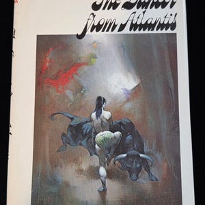 May include: A paperback book titled "The Dancer from Atlantis" with a cover illustration. The artwork depicts a figure between two black bulls. The author's name, Poul Anderson, is at the bottom. The book has a white background.