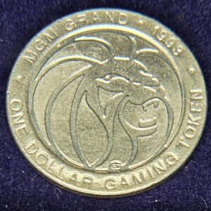 May include: A silver-colored MGM Grand one-dollar gaming token from 1998. The token features a lion's head in the center, surrounded by the words "MGM GRAND" and "ONE DOLLAR GAMING TOKEN".