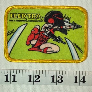 May include: Rectangular patch featuring the character Elektra from Marvel Comics. The patch has a green background with a yellow border. Elektra is depicted in a red outfit, holding a weapon. The text "ELEKTRA" and copyright information are visible.