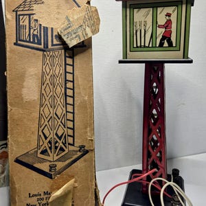 May include: Vintage toy train signal tower with a red and black metal frame and a red brick roof. The signal box features a green and white window with a figure inside. The original cardboard box is included.
