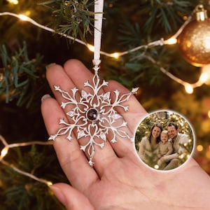May include: Silver snowflake Christmas ornament with a dark center stone and clear accents. The ornament is hanging from a white ribbon. A small, circular photo of a family is in the background. Christmas decor.