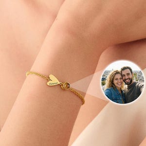 May include: Gold-colored bracelet with a heart-shaped charm. The charm has the word "Forever" engraved on it. The bracelet is on a person's wrist, with a small photo of a couple in the background.