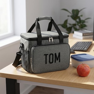 May include: A rectangular grey insulated lunch bag with black trim and handles. The front pocket features the name TOM in bold black letters. The bag includes a top handle, a detachable shoulder strap, and a side mesh pocket for storage.