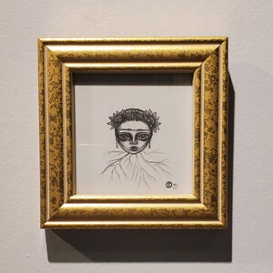May include: A framed pencil drawing of a stylized face with large eyes, a floral crown, and a detailed collar. The artwork is in a gold-colored frame with a textured finish, mounted on a light gray wall.