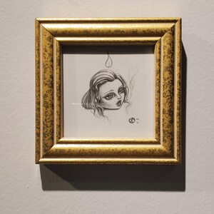 May include: A framed black and white drawing of a woman with a single teardrop above her head. The drawing is in a gold frame with a textured surface.