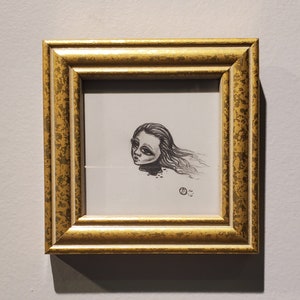 May include: A framed black and white drawing of a woman's head with long hair, the drawing is in a gold frame with a textured surface.