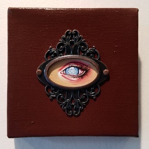 May include: A small, framed painting of a blue eye with a red eyelid. The frame is ornate and made of metal with a brown background.