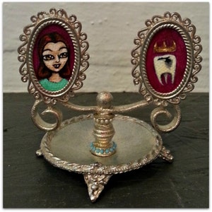 May include: A silver decorative stand with two round mirrors. The mirrors have ornate frames and feature painted portraits. One portrait is of a woman with red hair and the other is of a tooth with a crown.