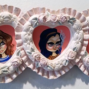 May include: Three heart-shaped frames with pink and white scalloped edges, each containing a painted portrait of a girl with large eyes. The frames are decorated with pink roses and green leaves.