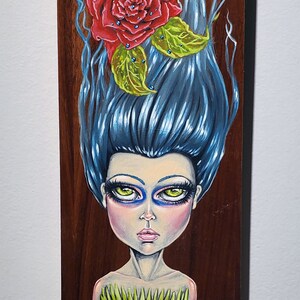 May include: A painting of a woman with blue hair and a red rose on her head. She is wearing a green dress with a leafy design. The background is a dark brown wood.