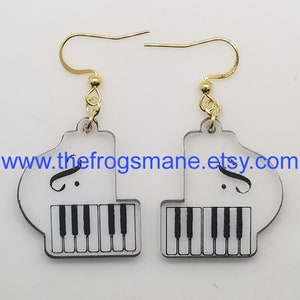 May include: A pair of white acrylic earrings shaped like a grand piano with black keys and a treble clef. The earrings have gold hooks.