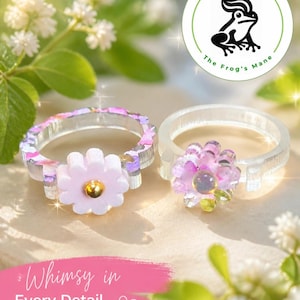 May include: Two clear resin rings with floral embellishments. One ring features a pink flower with a gold center, the other a purple flower. The rings are displayed on a surface with the text "Whimsy in Every Detail". The Frog's Mane logo is also visible.